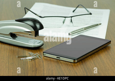 Still-life photography on office desk with stapler and office staples next to mobile phone in front of paper with glasses and pen - blurred background Stock Photo