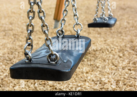Close-up view of a still child's swing in black plastic in a wood chips covered playground with chrome chains. Stock Photo
