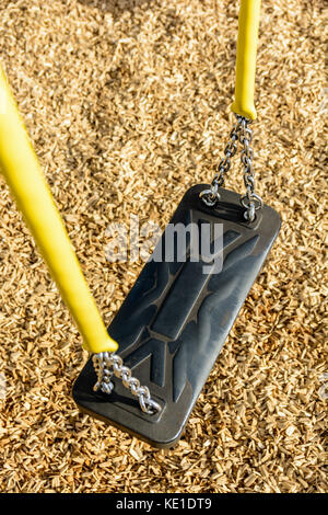 A still child's swing in black plastic in a wood chips covered playground with chrome chains in a yellow plastic sleeve. Stock Photo
