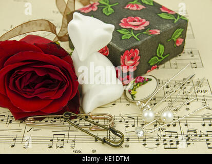 Still life with red rose and angel among several hairpins, gift boxes and buttons on the background of paper with notes Stock Photo