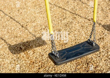 A still child's swing in black plastic in a wood chips covered playground with chrome chains in a yellow plastic sleeve and drop shadow. Stock Photo