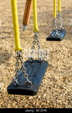 A still child's swing in black plastic in a wood chips covered playground with chrome chains in a yellow plastic sleeve. Stock Photo