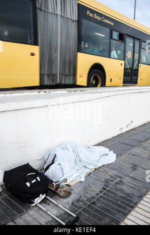 homeless person with skateboard and wheeled suitcase sleeping near busy road in Spain Stock Photo