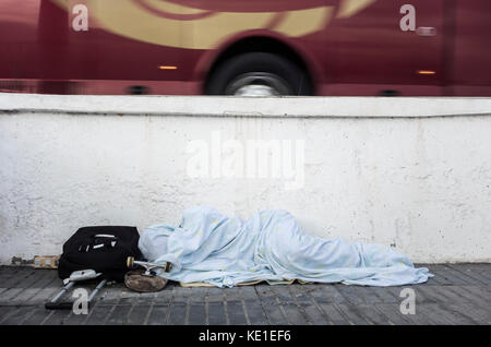 homeless person with skateboard and wheeled suitcase sleeping near busy road in Spain Stock Photo