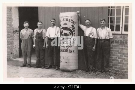 Hop pickers or workers with hops packed in a sack or 'poke', possibly standing outside the hop kiln - This photo depicts workers on the Kingston Bagpuize estate, near Abingdon, Oxfordshire (at this time it was in the county of Berkshire). The hop garden, was owned by the Berkshire Hop Company who bought the gardens in 1936 from the former owner of the Estate, Edward Strauss. The 'poke' has the white horse logo and states at the top 'successors of E.A. Strauss. Stock Photo