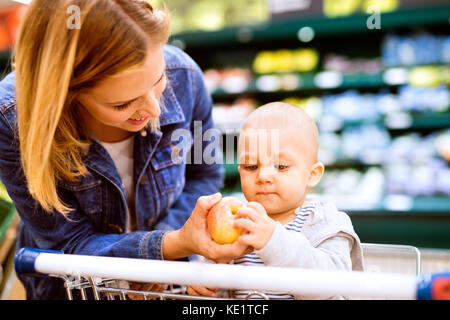 Beautiful young mother with her little baby boy at the supermarket, shopping. Woman giving her son an apple. Close up. Stock Photo