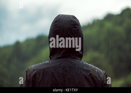 Rear view of hooded man standing in rain on cold cloudy autumn day getting drenched Stock Photo