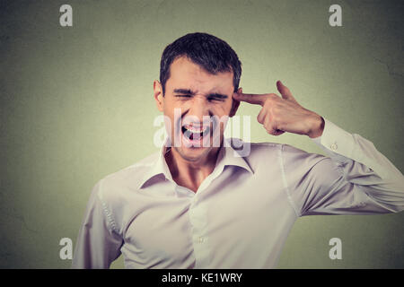 Headshot portrait young stressed man committing suicide with finger gun gesture isolated on grey wall background. Human emotion face expression. Overw Stock Photo