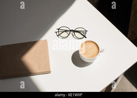 top view of cup of coffee, eyeglasses and notepad on white table with sunlight Stock Photo
