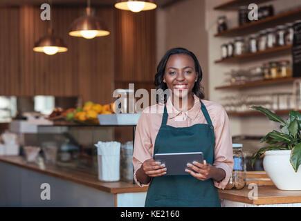 Portrait of a smiling young African female entrepreneur in an apron using a digital tablet while standing in her trendy cafe Stock Photo