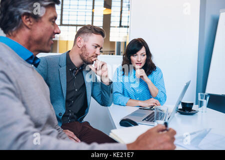 Three work colleagues talking and using a laptop together while sitting at a table in a meeting room in a modern office Stock Photo