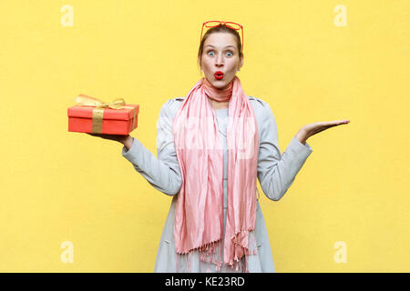 Hand on sides and holding christmas box. Woman looking at camera with shocked face and open mouth. Studio shot, isolated on yellow wall Stock Photo