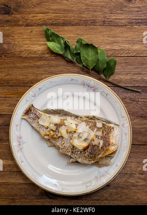 Oven cooked whole fish, with garlic and onion sliced on top, on a white porcelain decorative plate, on a light wooden background, with a bay leaf bran Stock Photo