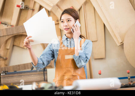 Young smiling female woodworker with blueprint talking on cellphone Stock Photo