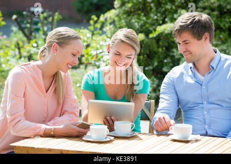 Group Of Smiling Friends Looking At Digital Tablet With Cup Of Coffee On Table Stock Photo