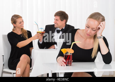 Depressed Woman Sitting In Restaurant In Front Of Couple Toasting Glass Of Cocktails Stock Photo