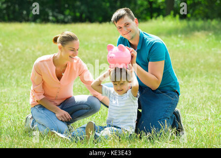 Happy Parents With Their Son Holding Pink Piggy Bank On His Head In The Park Stock Photo