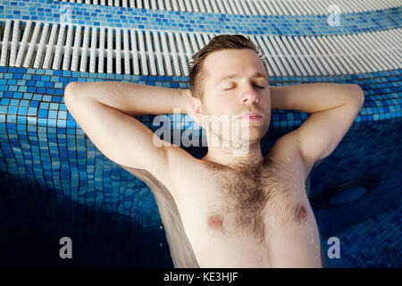 The man is resting on the edge of the pool. Hands are behind the head. Stock Photo