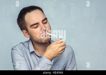Young man lighting cigarette. Businessman in a shirt he lights a cigarette from the flame of the lighter on a blue background. Stock Photo