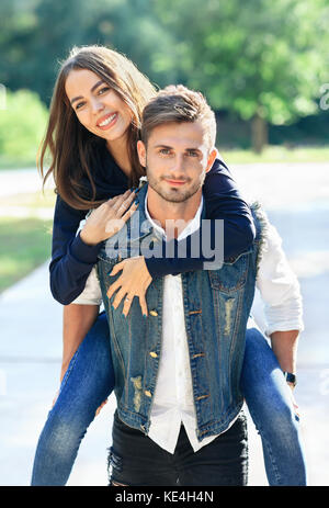 Girl rides on back of guy, smiling at camera outdoors. Loving couple piggyback in nature in park. Two young people having fun as children in the open  Stock Photo