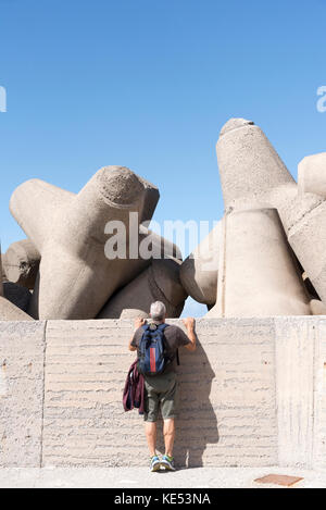 Man on tiptoe inspecting the man made concrete blocks, Tetrapods, which act as seawall defence around a harbour wall. Crete, Greece, 2017 Stock Photo