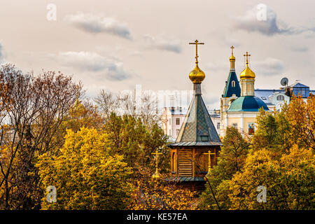 View of a small wooden church in the big city in the golden autumn season. Gray clouds in the beige color sky. Stock Photo