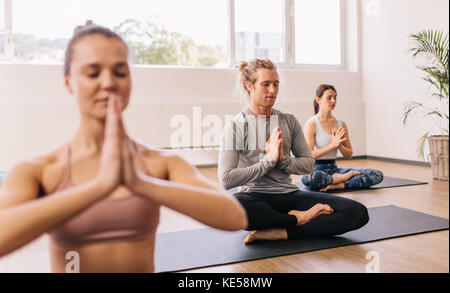People meditating while sitting in room. Young man practicing yoga in gym class with people sitting around. Stock Photo