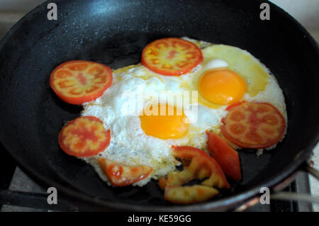 Two fried eggs in black frying pan and slices tomato. Process of cooking. Stock Photo