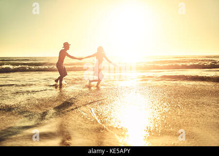 Young couple holding hands, walking in sunny summer sunset ocean beach surf Stock Photo