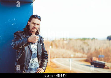 Male biker in a leather jacket and bandana puts on sunglasses and looking at the camera. Motorcyclist in leather jacket puts on sunglasses. Stock Photo