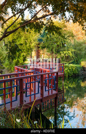 A red and blue zig-zag bridge crossing a calm pond to a gazebo in a park like setting in fall. Stock Photo