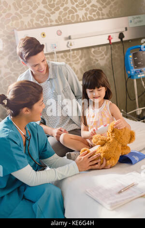 Female nurse and girl patient using digital thermometer on teddy bear in hospital room Stock Photo