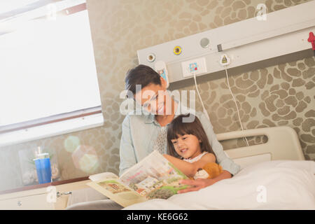Mother reading book with daughter patient in hospital room Stock Photo