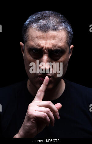 Furious mature man with aggressive look making silence sign in violent and threatening way. Black background / rage shh shhh shut up scary angry Stock Photo