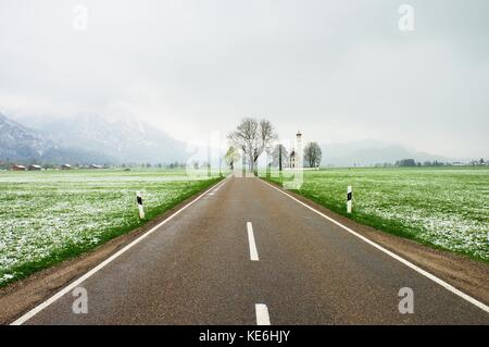 Straight  asphalt way between meadows. April weather. Wet spring snow in already fresh green grass bellow mountains. Stock Photo