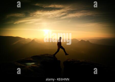 Crazy hiker in black is jumping between rocky peaks. Happy man. Wonderful daybreak in rocky mountains, heavy orange mist in deep valley. Miracle of na Stock Photo
