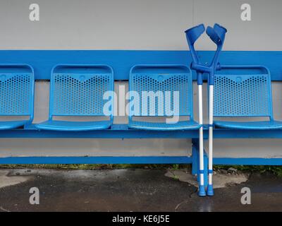 Medical crutch at blue metal seats on outdoor stadium players bench, rusty chairs with old paint below transparent plastic roof. Stock Photo