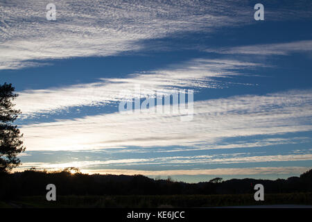 A blue sky in late afternoon with the setting sun highlighting striped clouds and framed by landscape and trees. Stock Photo