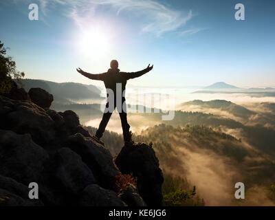 Sunny fall morning. Happy hiker with raised hands in air stand on rock above pine forest. View over misty and foggy morning valley to Sun. Stock Photo