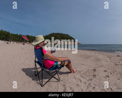 Woman reads her Kindle on the beach, Black Brook Cove, Cape Breton Highlands National Park, Cape Breton Island, Nova Scotia, Canada. Stock Photo