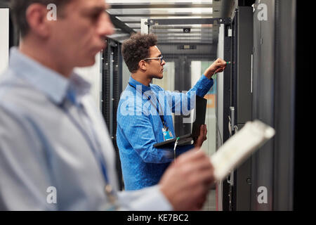 Male IT technicians with clipboard and laptop working at panels in server room Stock Photo