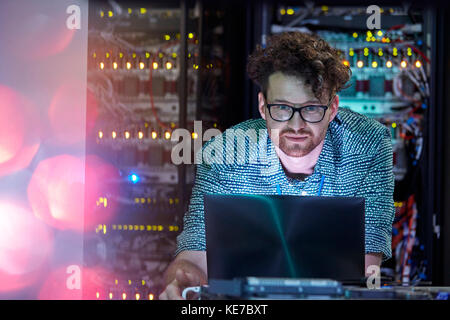 Focused male IT technician working at laptop in dark server room Stock Photo