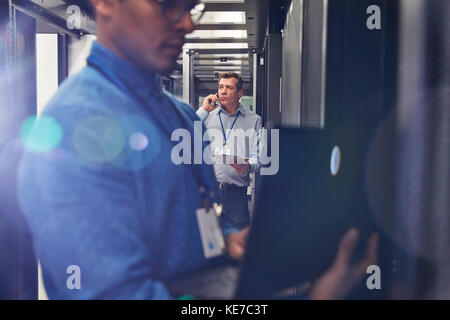 Male IT technicians using laptop and talking on cell phone in server room Stock Photo