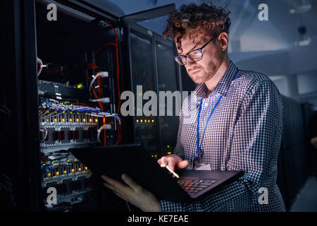 Focused male IT technician working at laptop in dark server room Stock Photo