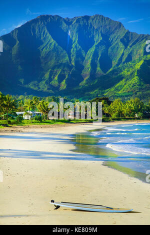 A surfboard sits on the beach at the water's edge with rugged green mountains and lush foliage on the island of Kauai Stock Photo