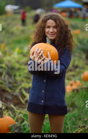 Portrait of a young smiling woman holding a pumpkin at a farm pumpking patch, British Columbia, Canada, autumn harvest Stock Photo