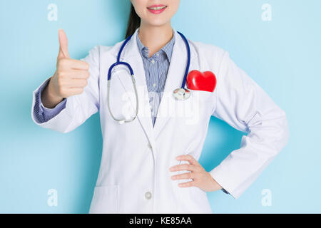 smiling young doctor woman making thumb up gesture showing good cardiolog healthy care concept standing in blue wall background. Stock Photo