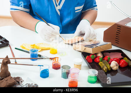 Close-up of a woman a pastry-shop painter with a brush of milk chocolate with edible paint, on a white table there is chocolate bread, strawberries, g Stock Photo