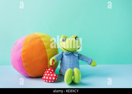 Kids Toys Small Plush Frog Holding Red Heart Multicolored Textile Soft Ball on Blue Green Background. Banner Placeholder Charity Nursery Hospital Copy Stock Photo