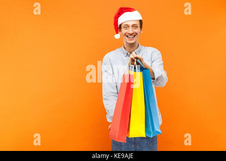 Young adult joyful man in christmas red hat, toothy smiling, showing colorful packets at camera. Studio shot on orange background Stock Photo